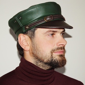 Op de afbeelding: Een groene leren pet met een bruin vizier en riem, met een goudkleurige gesp. De pet heeft een gestructureerd ontwerp en wordt schuin gedragen. De pet is een stijlvol accessoire.