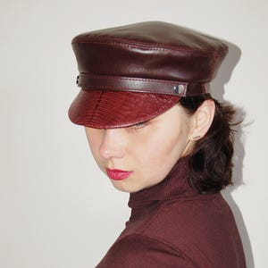 May include: A burgundy leather baker boy hat with a textured visor and a strap detail. The hat has a rounded crown and a structured brim. The hat is a stylish accessory.