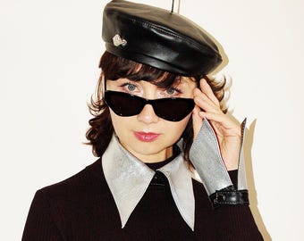 May include: A black leather beret with a small silver-toned accent, black cat-eye sunglasses, a detachable metallic silver collar, and a matching metallic silver cuff with a black leather strap and buckle, worn over a black ribbed long-sleeve top.