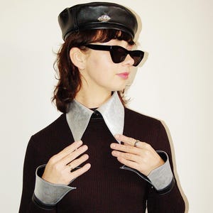 Peut inclure: Un béret en cuir noir orné d'un bijou, associé à des lunettes de soleil noires et un col et des poignets argentés. Le béret est un accessoire classique, tandis que les accents argentés ajoutent une touche moderne à la tenue. L'ensemble est élégant et chic.