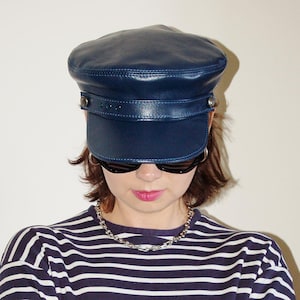 Op de afbeelding: Een marineblauwe leren kapiteinshoed met een gestructureerde klep en decoratieve band. De hoed heeft zilverkleurige metalen accenten. De persoon draagt een zwarte zonnebril en een gestreept marineblauw en wit shirt.