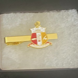 May include: A gold tie bar with a white, red, and gold crest featuring a bird and the text "Alpha Kappa Alpha"