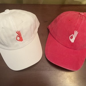 May include: Two baseball caps, one white and one pink, with a white embroidered OK hand gesture on each.