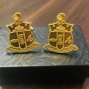May include: A pair of gold cufflinks in a black box. The cufflinks feature a shield design with a Greek letter and other symbols.