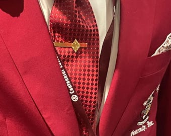 Op de afbeelding: Een rood colbert met een rood-witte geblokte das, een gouden dasclip en een zwarte bloemspeld. Het colbert heeft een wit zakdoekje en een klein Amerikaans vlagpinnetje. Het colbert heeft ook een geborduurd embleem met de tekst "Fraternity, Inc."