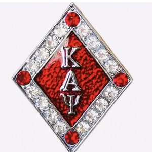May include: A red diamond-shaped pin with a silver border and white rhinestones. The center of the pin features the letters "ΚΑΨ" in silver.