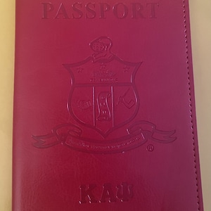 May include: A red leather passport holder with the word "PASSPORT" embossed on the front. The holder features a crest with the letters "KA".