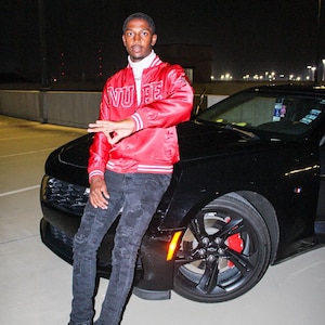 May include: A young person wearing a red satin bomber jacket with the word "NUPE" embroidered on the front, black jeans, and black and white sneakers. The person is sitting on the hood of a black car.