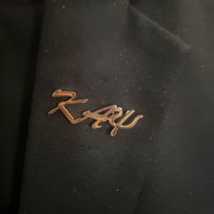 May include: A gold-colored pin with the word "Ray" written in cursive script.
