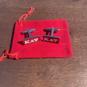 May include: A pair of silver cufflinks with red enamel and the letters "KAY" engraved on each cufflink. The cufflinks are in a red velvet pouch.