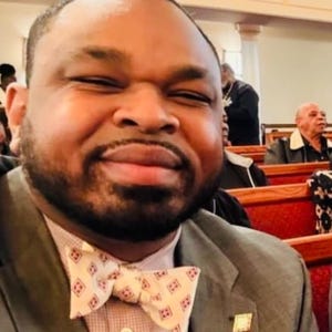 May include: A man wearing a gray suit jacket with a pink shirt and a patterned bow tie. He is smiling and looking at the camera.