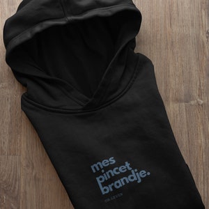 Op de afbeelding: Zwart hoodie met de tekst "mes pincet brandje." en de woorden "OK-LEVEN" eronder gedrukt.