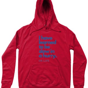 Op de afbeelding: Rood hoodie met de tekst "I have learned to be slow in a hurry. OR-LIFE" in blauw gedrukt.