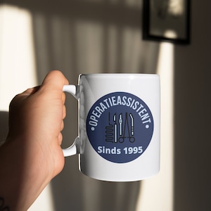 Peut inclure: Une tasse en céramique blanche avec un motif circulaire bleu. Le cercle présente un graphique d'instruments chirurgicaux et le texte "OPERATIEASSISTENT Sinds 1995".
