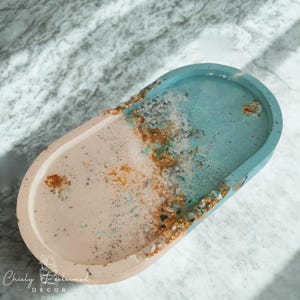 May include: An oval decorative tray with a two-tone design. The tray features a light pink and teal color scheme, with gold and silver accents along the edges. The tray is made of resin and has a smooth, glossy finish. The tray is labeled "Chicly Reclaimed Decor."