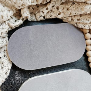 May include: A gray, oval-shaped decorative tray with a textured surface. The tray is made of a soft material and is displayed on a dark surface. The image also shows a cream-colored lace and wooden beads.