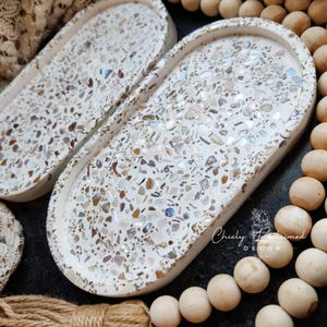 May include: Two oval-shaped decorative trays with a white base and a mosaic of iridescent and neutral-toned chips. The trays have a raised edge and are set against a dark background with wooden beads and rope.
