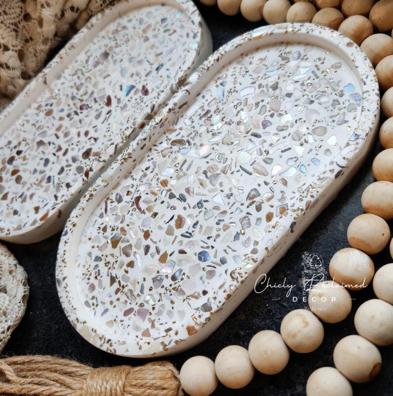 May include: Two oval-shaped decorative trays with a white base and a mosaic of iridescent and neutral-toned chips. The trays have a raised edge and are set against a dark background with wooden beads and rope.