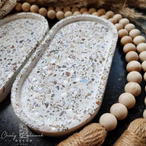 May include: Two oval decorative trays with a white terrazzo design, featuring iridescent and gold accents. The trays are surrounded by wooden beads and a jute tassel. The trays are from Chicly Reclaimed Decor.