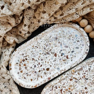 May include: Oval decorative tray with a terrazzo design. The tray features a white base with embedded chips of various colors, including brown, beige, and gray. The tray is part of a home decor collection.