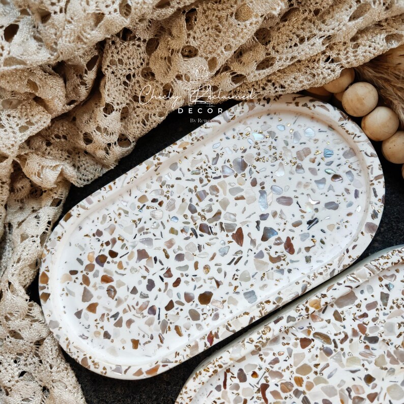 May include: Oval decorative tray with a terrazzo design. The tray features a white base with embedded chips of various colors, including brown, beige, and gray. The tray is part of a home decor collection.
