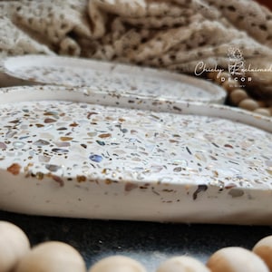 May include: White decorative tray with a speckled design. The tray's surface is covered in a mosaic of small, colorful pieces, including gold, brown, and iridescent elements. The tray has a raised edge and a smooth, rounded shape. The text "Chicly Reclaimed Decor" is visible.