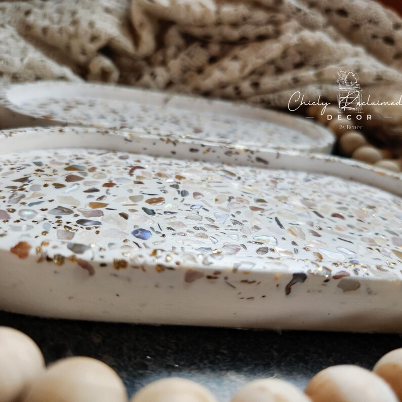 May include: White decorative tray with a speckled design. The tray's surface is covered in a mosaic of small, colorful pieces, including gold, brown, and iridescent elements. The tray has a raised edge and a smooth, rounded shape. The text "Chicly Reclaimed Decor" is visible.