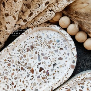 May include: Oval decorative tray with a terrazzo design in shades of white, brown, and iridescent accents. The tray is part of a home decor collection. The tray has a smooth surface and a slightly raised edge. The tray is next to a beige lace fabric.
