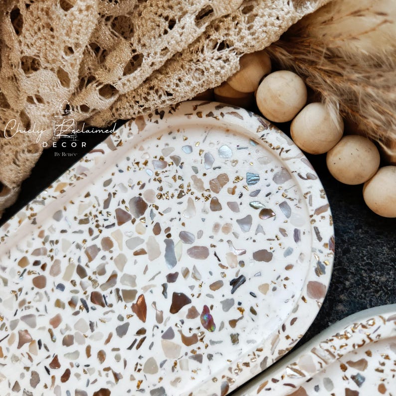 May include: Oval decorative tray with a terrazzo design in shades of white, brown, and iridescent accents. The tray is part of a home decor collection. The tray has a smooth surface and a slightly raised edge. The tray is next to a beige lace fabric.