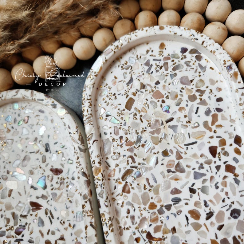 May include: Two white decorative trays with a terrazzo design. The trays are oval-shaped with a raised edge and are filled with a mosaic of colorful chips. The trays are decorated with gold accents and the text "Chicly Reclaimed Decor" is visible.