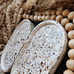 May include: Two white, oval-shaped decorative trays with a terrazzo design. The trays are filled with a mosaic of small, colorful stones and flecks of gold. The trays are labeled "Chicly Reclaimed Decor By Renee".