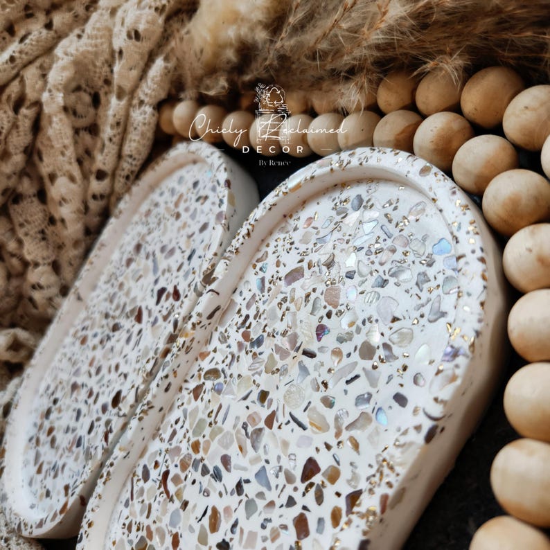 May include: Two white, oval-shaped decorative trays with a terrazzo design. The trays are filled with a mosaic of small, colorful stones and flecks of gold. The trays are labeled "Chicly Reclaimed Decor By Renee".