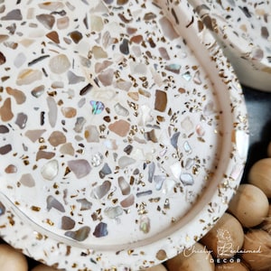 May include: A close-up of a white decorative tray with a terrazzo design. The tray is speckled with various colors of stone, including beige, brown, and gray, with gold accents. The tray has a slightly raised edge and a smooth surface. The overall theme is home decor.