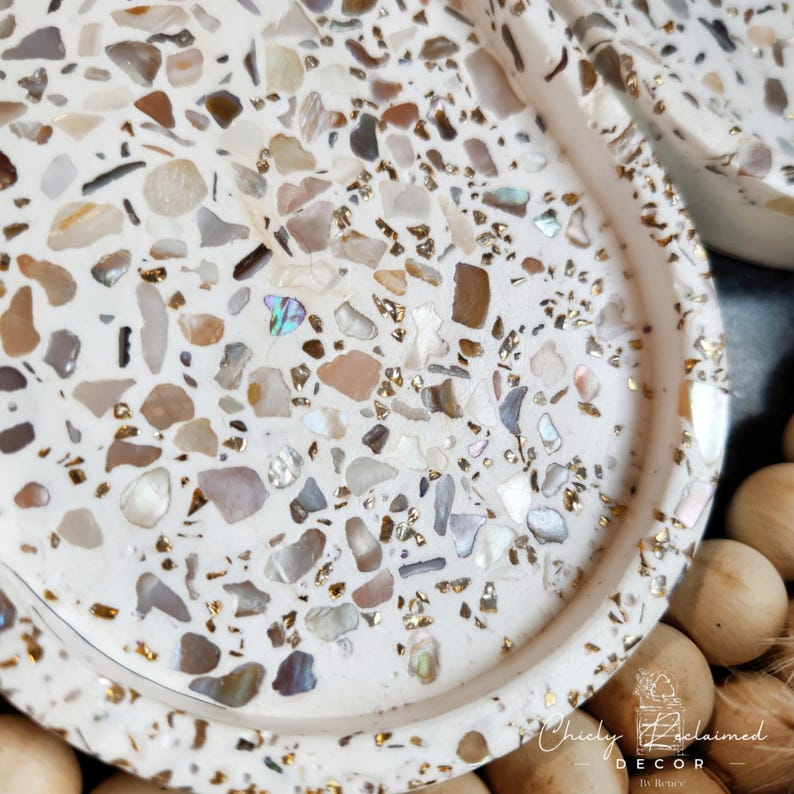 May include: A close-up of a white decorative tray with a terrazzo design. The tray is speckled with various colors of stone, including beige, brown, and gray, with gold accents. The tray has a slightly raised edge and a smooth surface. The overall theme is home decor.