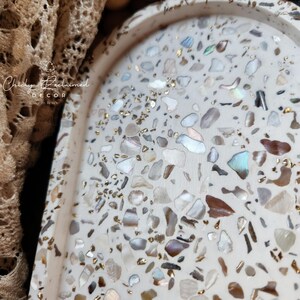 May include: A decorative, off-white tray with a terrazzo design. The tray is speckled with various shapes of iridescent, brown, and gray stones, and gold flecks. The tray has a curved, oval shape. The text "Chicky Reclaimed Decor" is visible.