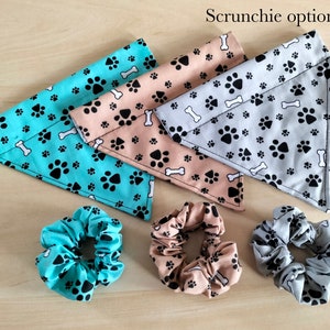 Op de afbeelding: Drie hondenbandanas in turquoise, beige en grijs met een poot- en botpatroon. De tekst 'Scrunchie optional' is zichtbaar bovenaan de afbeelding. Er zijn ook drie bijpassende scrunchies te zien op de afbeelding.