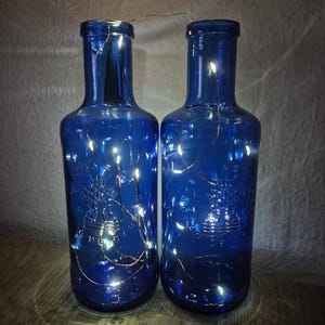 Puede incluir: Dos botellas de vidrio azul con luces de cadena en su interior. Las botellas tienen un diseño en relieve y están iluminadas por pequeñas luces LED blancas cálidas. Las botellas están de pie, creando un brillo ambiental suave.