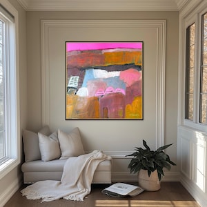 May include: Abstract painting with a black frame. The artwork features a vibrant color palette of pink, orange, blue, and yellow. The painting is displayed above a bench with pillows and a blanket, next to a window and a potted plant.