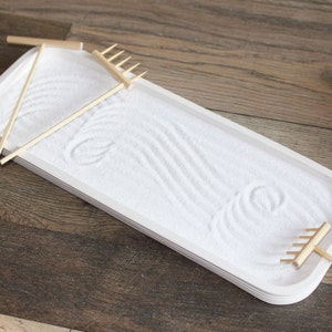 May include: A white rectangular tray with a layer of white sand. The tray has two wooden rakes and a wooden stick for creating patterns in the sand.