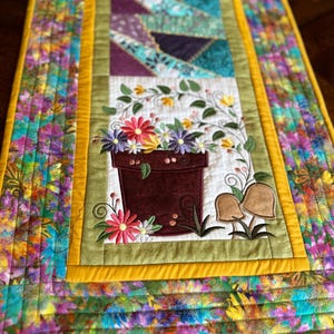 May include: A quilted table runner with a floral design. The runner features a brown flower pot with colorful flowers and green vines. The runner has a yellow border and a multi-colored floral pattern.