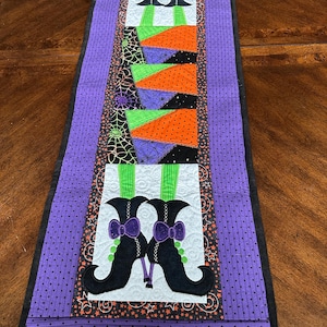 May include: A Halloween-themed table runner featuring a patchwork design. The runner showcases witch legs and boots in black, green, orange, and purple. The border is a dotted purple fabric with a black edge. The design includes bows, stars, and spiderwebs.