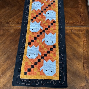 May include: A Halloween-themed table runner featuring a repeating pattern of white ghost figures on an orange and yellow background. The ghosts are set against a black border with decorative stitching. The runner is approximately 60 inches long.