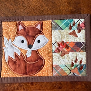 May include: A quilted coaster featuring a cartoon fox with a white belly and tail, set against an orange dotted background. The right side has plaid fabric with embroidered paw prints, all framed with a brown border.