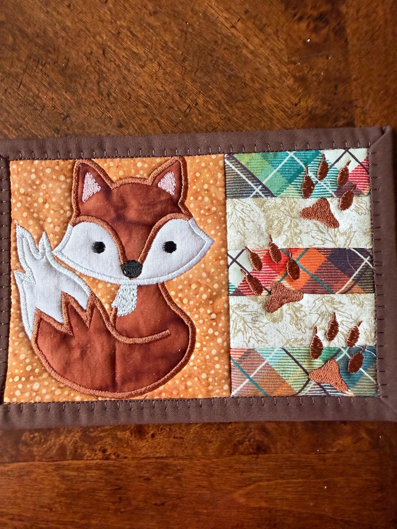 May include: A quilted coaster featuring a cartoon fox with a white belly and tail, set against an orange dotted background. The right side has plaid fabric with embroidered paw prints, all framed with a brown border.