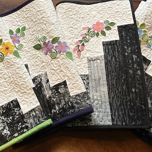 May include: Four quilted table runners with a black and white woodgrain design and embroidered floral accents. The runners have different colored flowers: yellow, purple, pink, and blue.