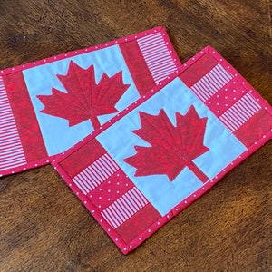 May include: Two red and white Canadian flag-themed coasters with a white background and a red maple leaf design. The coasters have a white polka dot border and a red and white striped border.