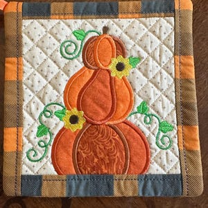 May include: A quilted pot holder with a brown, black and orange plaid border. The centre features an embroidered design of three stacked pumpkins with green vines and yellow sunflowers.