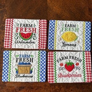May include: Four quilted fabric coasters with embroidered designs. Each coaster features the words "Farm Fresh" and a different fruit: watermelon, lemons, blueberries, and strawberries. The coasters have gingham borders in red and blue.