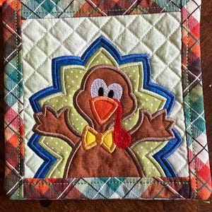 May include: Square quilted potholder with a Thanksgiving turkey design. The turkey is brown with an orange beak and red wattle, set against a green and blue starburst. The border is a plaid pattern in orange, green, and brown.