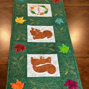 May include: A green table runner with a white background features a repeating pattern of squirrels, leaves, and wheat stalks. The word "Grateful" is embroidered in a wreath of leaves.